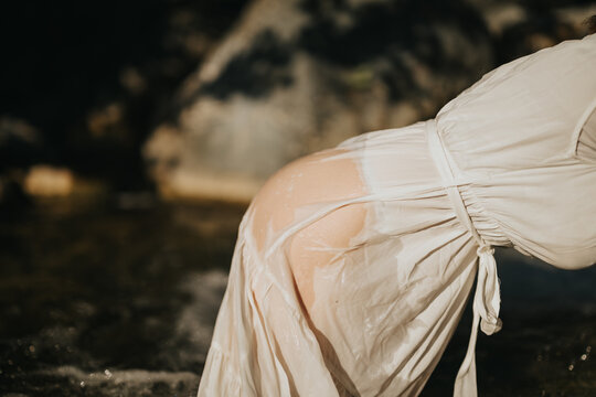 A woman is seen bending over in a white dress near a stream, with sunlight highlighting her wet attire. The scene captures both nature and tranquility.