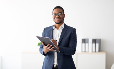 Happy young black male psychologist writing in clipboard, looking at camera and smiling in modern...