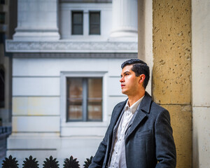A man in a dark coat leans against a textured stone wall, gazing thoughtfully into the distance. The background features an urban setting with classical architecture, including pillars and windows.