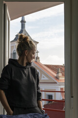 Blonde woman with hair up dressed in a tracksuit looks at the street through the window