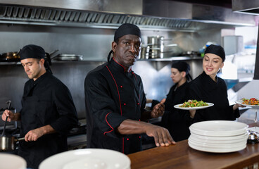 Portrait of confident African American man head chef posing in restaurant kitchen, team of chefs working behind
