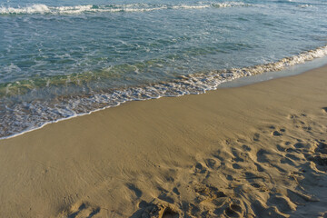 Gentle waves lap against the sandy shore under a bright blue sky at sunset by the ocean.
