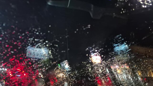 Rain at night on the side of the highway seen from the perspective of inside the car, you can see raindrops sticking to the car window. There is light from street lights, cars and other vehicles passi