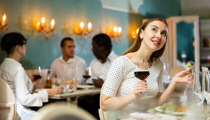 Positive young woman eating salad and drinking red wine in a cozy restaurant