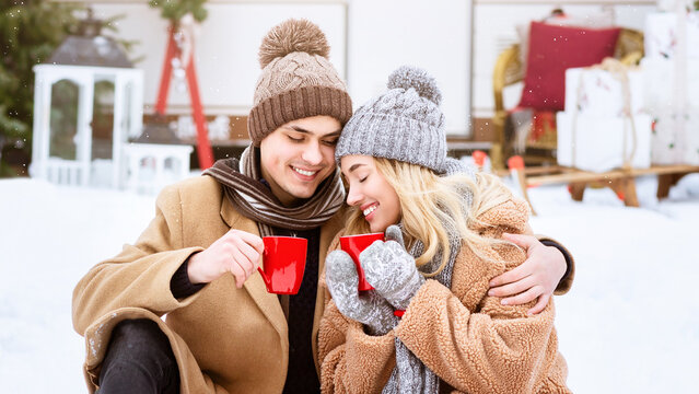 A young couple sits together in a snowy outdoor setting, sharing warm drinks from red mugs. They are bundled in cozy winter attire, radiating joy and warmth against the cold backdrop. - Powered by Adobe
