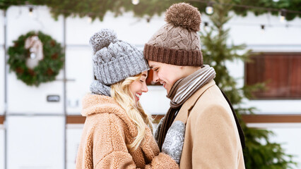 Two individuals share a warm embrace while dressed in cozy winter attire, surrounded by holiday decorations and winter greenery during a cheerful outdoor gathering.