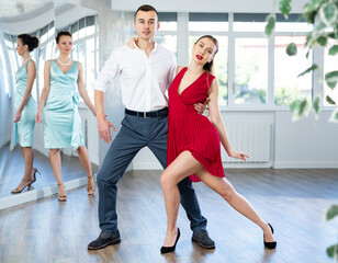 Positive young adult man in formal wear practicing impassioned samba with bright stylish female partner wearing red dress in dance class. Amateur dancing concept..