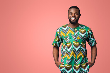 Handsome black man in traditional african clothes smiling over blue studio background, putting hands in pockets, copy space. Cheerful african american guy posing in ethnic colorful clothes