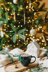Warm cup of chocolate with marshmallow, christmas ornaments, cozy knitted sweater and fir branches on background of stylish christmas tree with golden lights. Winter mood