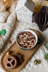 Warm cup of cocoa with marshmallows and chocolate cookies, christmas ornaments, cozy knitted sweater and fir branches on background of stylish christmas tree with golden lights. Winter mood