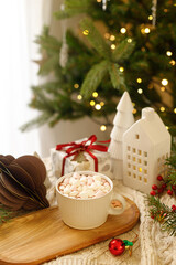 Warm cup of cocoa with marshmallows on wooden tray, christmas ornaments and gift on cozy knitted background against stylish christmas tree with lights. Winter mood. Merry Christmas