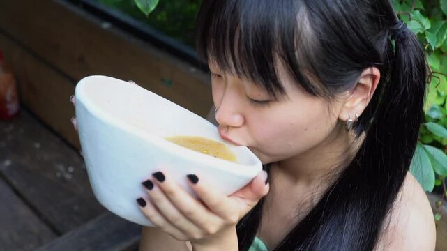 young asian woman drinking ramen from bowl in outdoor cafe, Korean woman scoops soup from bowl with ladle and slurps in restaurant on the street in the park