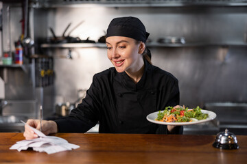 Young adult female chef cook checking orders and handing plate with meal through order station at restaurant kitchen