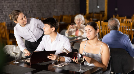 Young male and female colleagues having nice time in sparsely populated cozy restaurant. Waitress answers questions of visitors, helps to choose diet dish for late dinner in menu