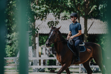 Fototapeta premium A young man confidently rides a horse during an outdoor equestrian training session, surrounded by lush greenery. He wears protective riding gear, showcasing his passion for horse riding and