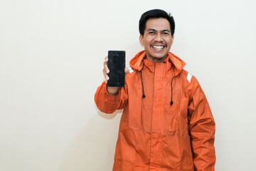 Smiling Southeast Asian man wearing orange raincoat looking at the camera and showing smartphone...
