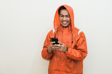 Smiling Southeast Asian man wearing orange raincoat holding a smart phone. Positive person....