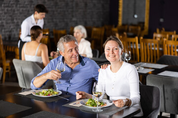 Adult woman and elderly man have dinner and drink wine together in restaurant