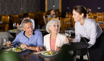 Adult woman waiter serving wine to couple of elderly man and woman in restaurant