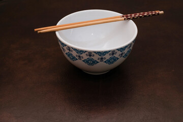 White bowl with blue pattern outside and wooden chopsticks on brown background, traditional wooden sticks for asian food.