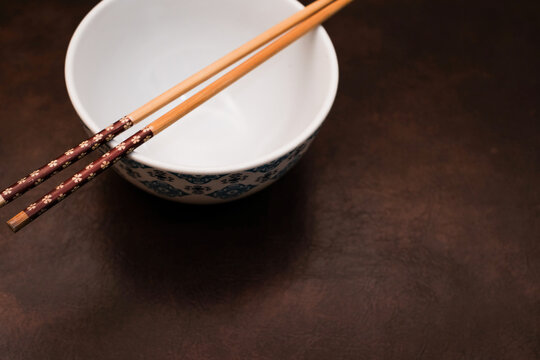 White bowl with blue pattern outside and wooden chopsticks on brown background, traditional wooden sticks for asian food.