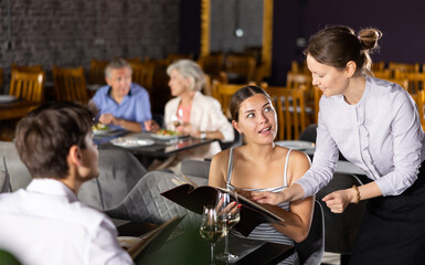 Adult female waiter in uniform takes order from young couple in restaurant