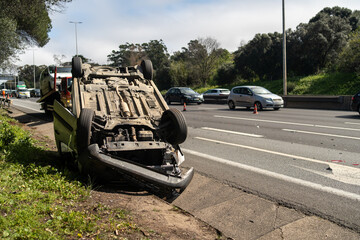 Damaged wrecked car after collision lying upside down on roadside. Busy city street with car rolled...