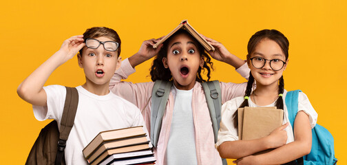 Education Concept. Set portrait of diverse schoolchildren posing looking at camera, holding stack of academic books and wearing backpack, yellow orange color studio background, panorama banner