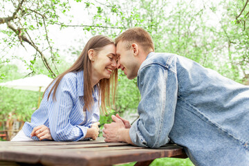young couple in love in denim clothes sitting in the garden at a wooden table and talking in the summer, guy and girl on a date talking and flirting in the park