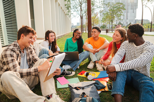 diverse group of happy university students sitting together on green campus lawn while studying reading and discussing with notebooks laptop and backpack during outdoor learning session - Powered by Adobe