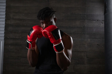 strong confident african american boxer in red gloves stands in a defensive stance near a punching bag, athlete in red gloves looks at the camera on a black background