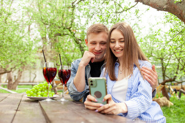 young couple in love in denim clothes sitting in the garden at a wooden table drinking wine with pork and using a smartphone in the summer, a guy and a girl on a picnic celebrating and taking selfies