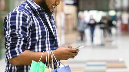 App With Discounts. Smiling Black Man With Shopping Bags Using Smartphone In Mall, Checking Sale...