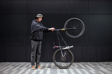 young hipster guy doing stunts on a bike in the city against the background of a black wall, street...