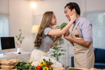 A man and a woman are in a kitchen, with the man holding a broccoli