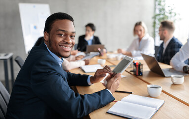 Happy African American Businessman Using Digital Tablet Computer Smiling To Camera Sitting At Corporate Meeting With Colleagues In Modern Office. Business Application Concept. Selective Focus