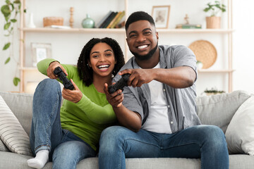 Cheerful black couple competing with each other in video games, holding joysticks and laughing,...