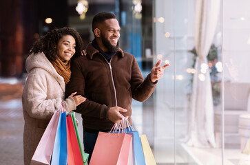 Sale And Consumerism Concept. Smiling african american couple looking and pointing at mall window, walking near shopping mall in the evening. Seasonal Sales And Discount, Purchase And Retail