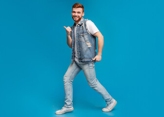 Cool millennial man dancing with thumbs up gestures on blue background, studio, copy space