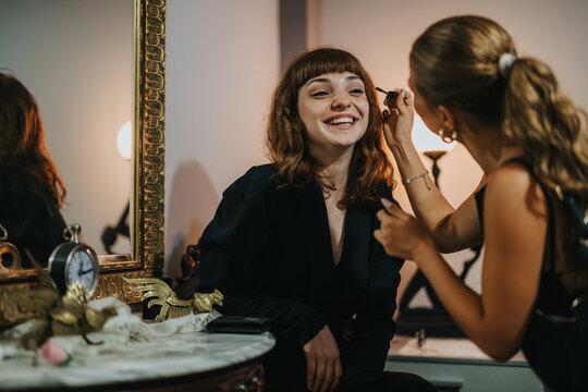 A joyful young woman smiles as a friend applies her makeup in a luxurious vintage room, showcasing warmth and friendship.