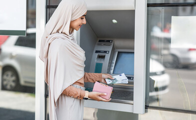 Concentrated cute young arab woman holds money withdrawn from ATM, muslim lady in hijab puts many euro in wallet near machine outside. Bank customer service, 24h service, easy banking operation