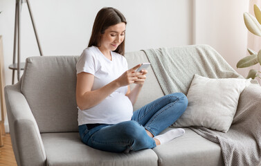 Obraz premium Pregnancy Tracker App. Smiling Young Pregnant Lady Using Smartphone At Home, Reading Baby Blog Or Browsing Internet, Happy Expectant Woman Relaxing With Cellphone On Couch In Living Room, Free Space