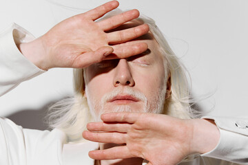 Portrait of a person with pale skin and white hair, shielding their face with hands against a light...