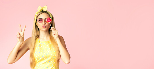 Sweet Life. Fashion portrait of girl in glasses holding lollipop, making peace sign, isolated over pink wall, copyspace