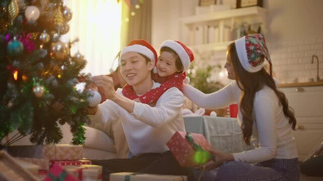 Happy family preparing New Year winter holidays celebration. Smiling young asian mother, father and son enjoy decorating xmas toy on christmas tree create festive mood atmosphere at home together.