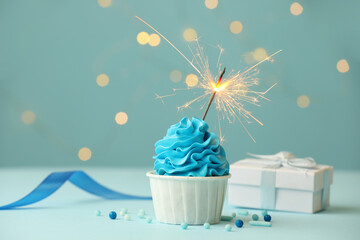 Birthday cupcake with burning sparkler and gift on light blue table against blurred lights