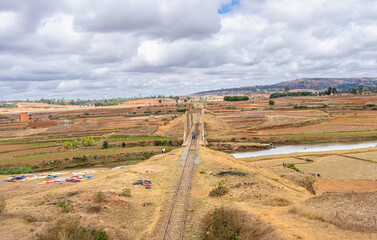 Scenic arched railway bridge crossing river in dry grassland with dramatic cloudy sky. Rural agricultural landscape with terraced fields and scattered trees, near Isalo National Park, Madagascar.