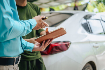 Asian clients engage with a car insurance agent beside a wrecked vehicle. They examine the policy details, assess the damage, and finalize paperwork related to the insurance claim and repairs.