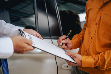 Asian clients engage with a car insurance agent beside a wrecked vehicle. They examine the policy...