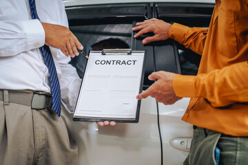 Asian clients engage with a car insurance agent beside a wrecked vehicle. They examine the policy details, assess the damage, and finalize paperwork related to the insurance claim and repairs.